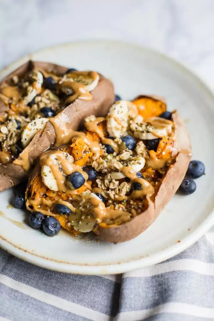 2 baked sweet potatoes on a plate filled with bananas, blueberries and granola and drizzled with peanut butter