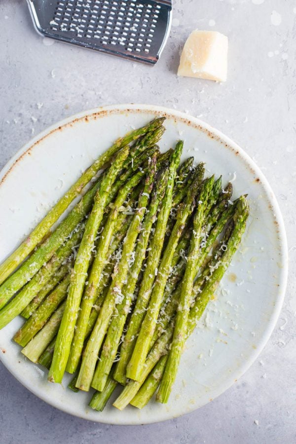 Plate full of cooked asparagus with parmesan cheese sprinkled on top and a cheese grate and cheese at the top of the photo