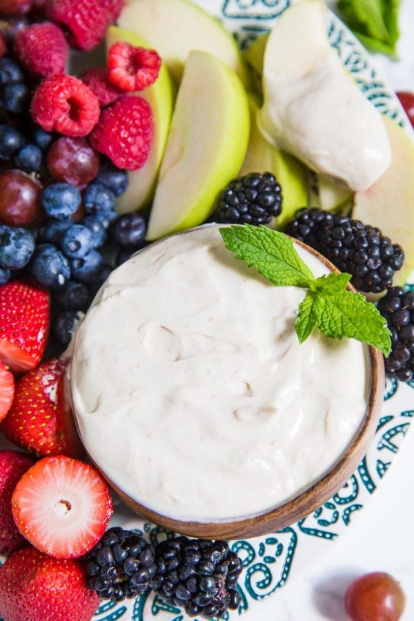 Small bowl of yogurt fruit dip on a platter that's surrounded by colorful fresh fruits including black berries, apples slices, strawberries, and blueberries