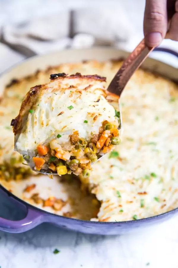 vegetarian pot pie with fresh mashed potatoes on top and fork indentions all in the top. Inside of a purple skillet and sitting on a marble board. A scoop is being taken out and you can see all the veggies on the inside of the pie