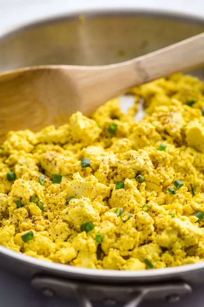 side shot of a stainless steel skillet with tofu scramble in it which consists of yellow tofu and green oninos
