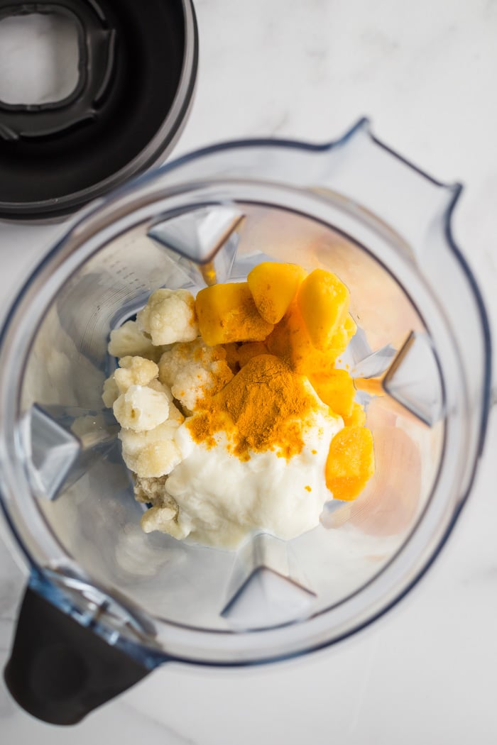 ingredients for a mango smoothie in a blender. top down shot. ingredients include cauliflower, mango, yogurt, and turmeric