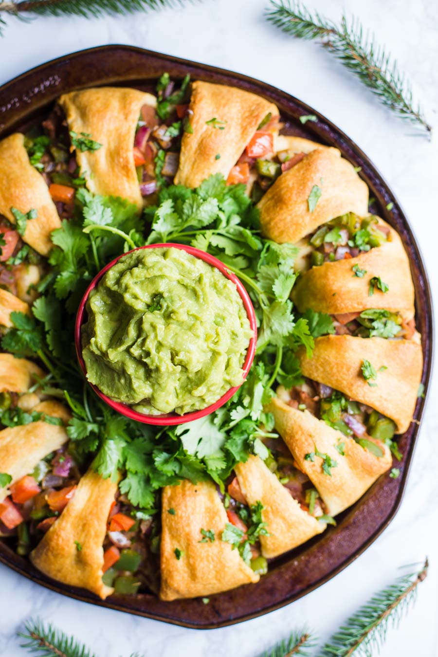 wreath of crescent rolls filled with taco fillings including veggies and beans. bowl of guacamole in the middle with fresh cilantro around the base of the bowl