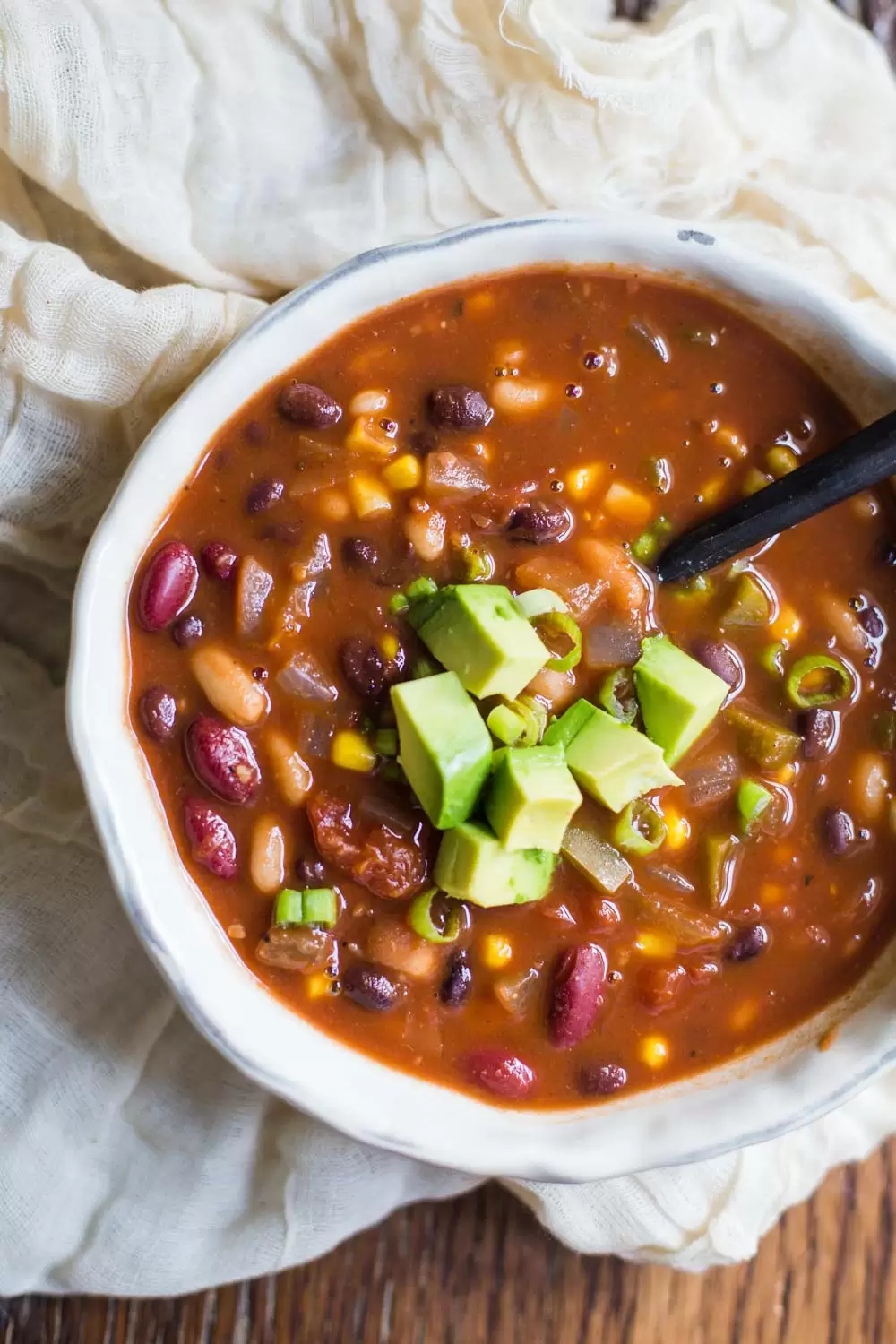 bowl of vegan chili topped with avocado on a wood table with a napkin