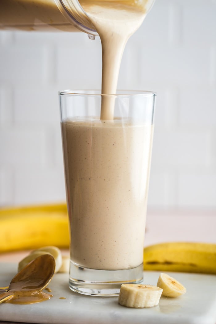 side shot of a peanut butter smoothie being poured into a clear glass. banana slices and fresh peanut butter around the glass. subway tile backdrop in the background