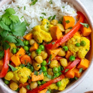 chickpea and sweet potato curry in a bowl that also has red pepper and peas in in served on white rice with cilantro on the side