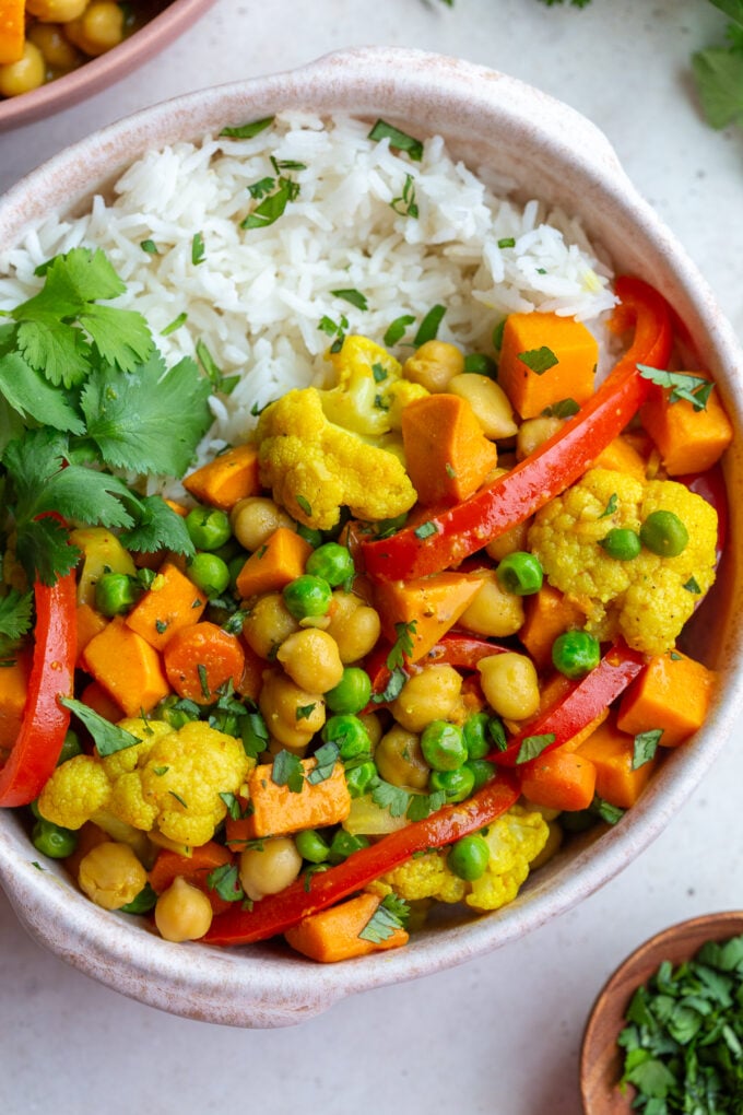 chickpea and sweet potato curry in a bowl that also has red pepper and peas in in served on white rice with cilantro on the side