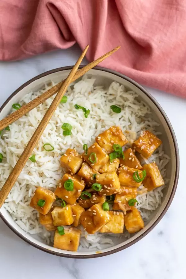 sauteed tofu on a bed of rice and topped with green onion and sesame seeds. on a marble board with a pink towel in the corner