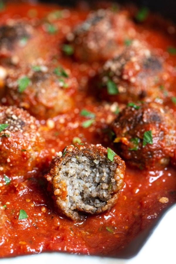 up close shot of vegetarian meatballs in a skillet with tomato sauce and sprinkled with vegan cheese and parsley. one with a bite taken out