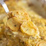scoop being taken out of the pan of golden scalloped potatoes topped with fresh herbs