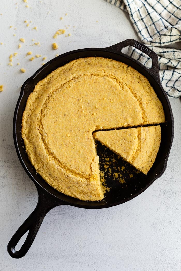 yellow cornbread in a black skillet with a slice cut out. a checkered towel in the background