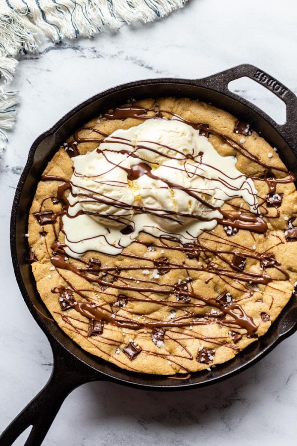 top down shot of black cast iron skillet with a skillet cookie in in covered in chocolate chunks, sea salt, and topped with 3 scoops of ice cream and drizzled with chocolate syrup. On a marble background with a stripped towel in the corner