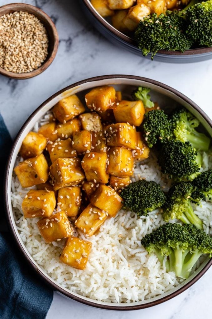 top down shot of orange sesame tofu cubes on a bed of rice with sauteed broccoli on the side and sprinkled with toasted sesame seeds. Sitting on a marble board with another bowl in the corner and a small bowl of sesame seeds on the side