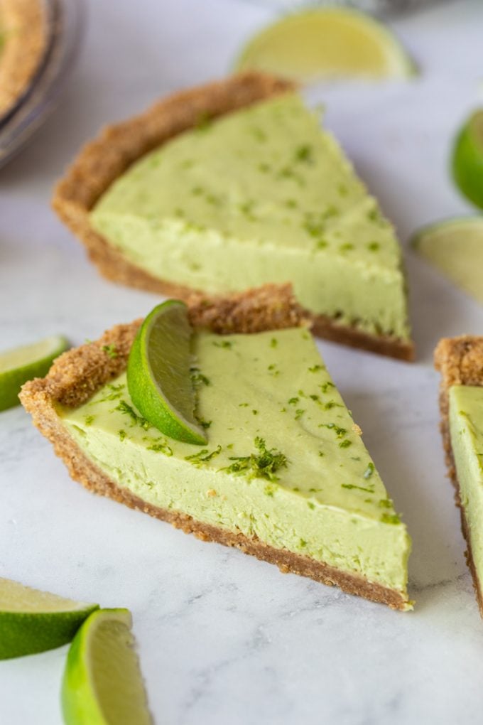 several slices of green key lime pie that's topped with lime zest and a lime wedge. Lime wedges all around the scene and everything sitting on a marble board