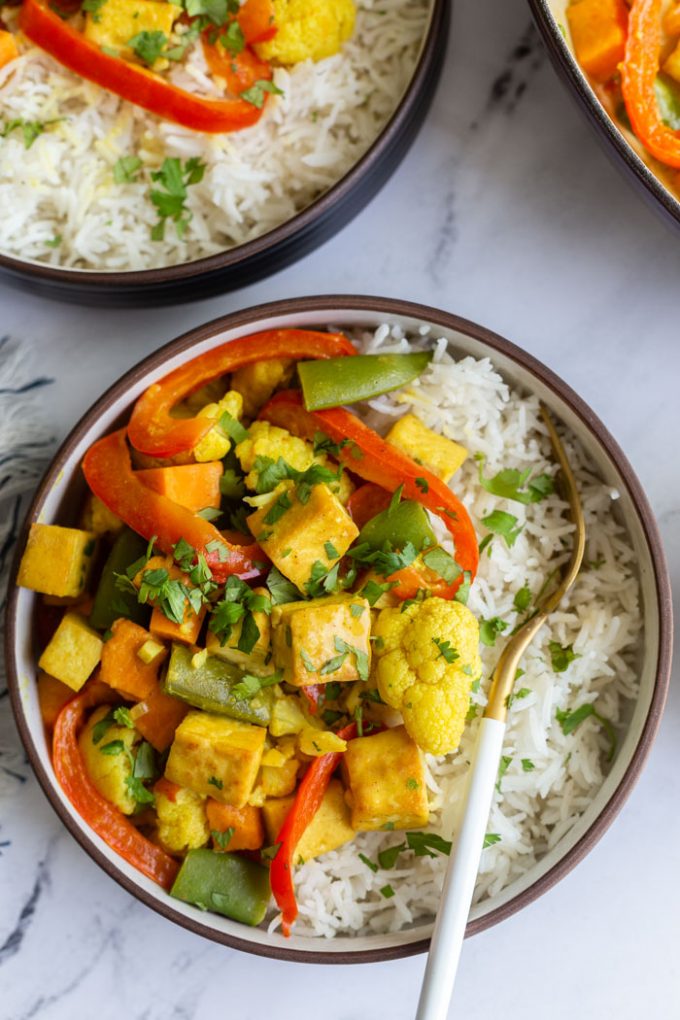 top down shot of a bowl of tofu curry that's yellow in color with red bell pepper, tofu, snap peas, and cilantro served over white rice