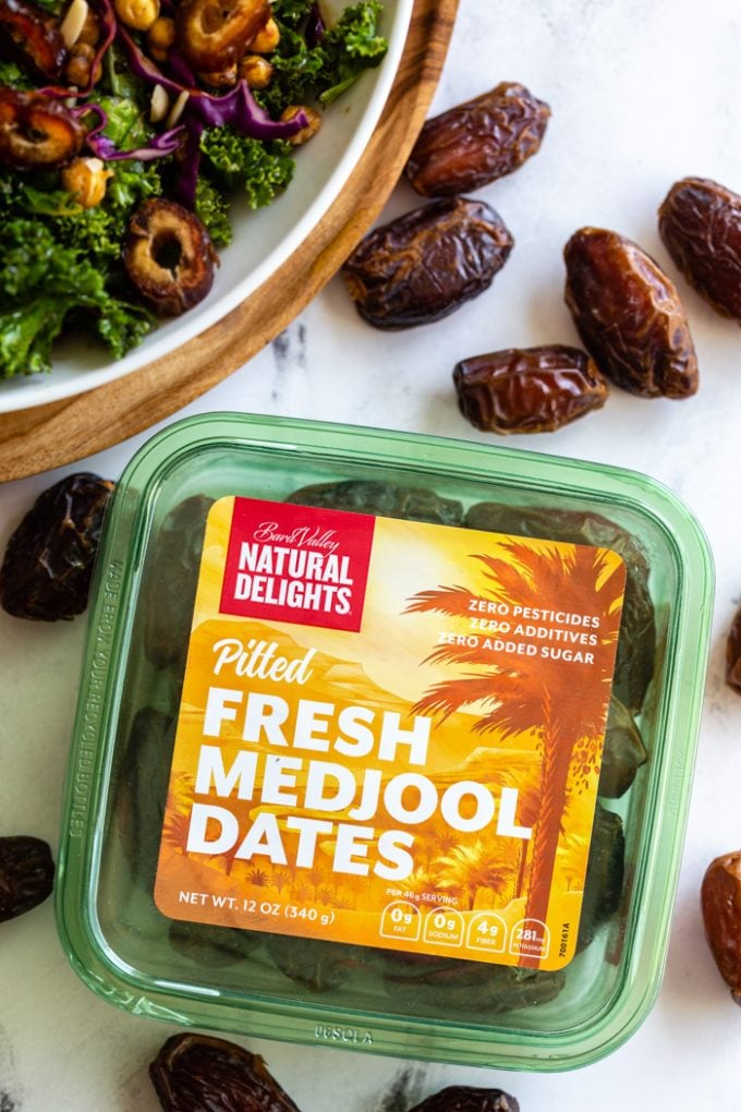 up close shot of a green box of Natural DelightsPitted Fresh Medjool Dates. a kale date salad is in the corner with fresh dates all around