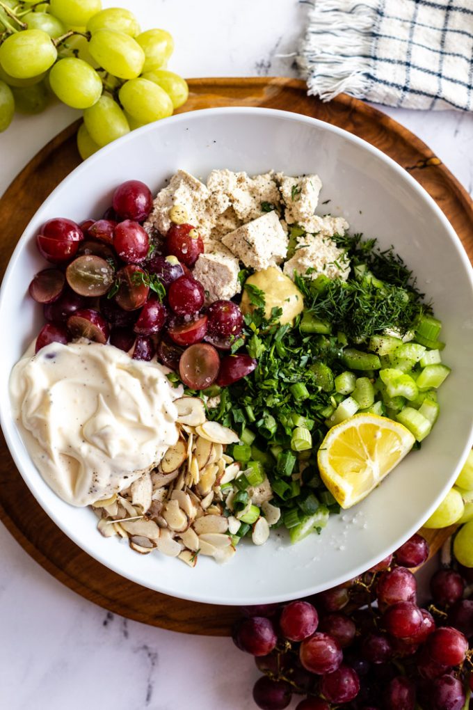 hvid skål fyldt med ingredienserne til denne tofu-kyllingesalat inklusive røde druer, mayo, nødder, grøn løg, dild, tofu, citron