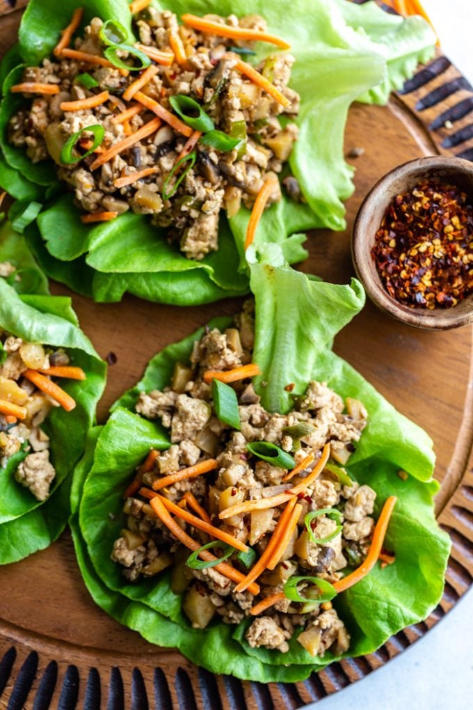 top down shot of vegan lettuce wraps filled with a tofu and mushroom mixture and topped with shredded carrots. all on a round wood board