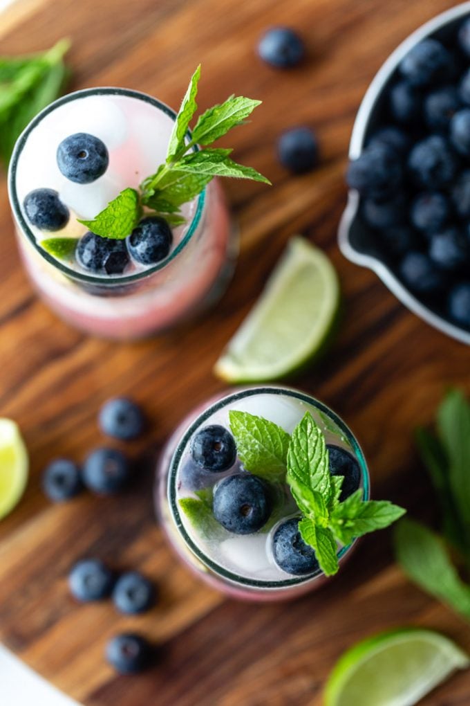 top down shot of 2 blueberry mojitos in clear glass with ice and fresh blueberries, mint, and lime garnishing the glass. Both sitting on a wood board with ingredients around