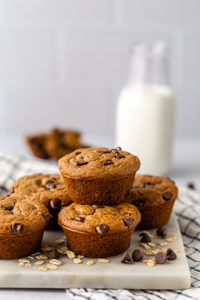 stack of chocolate oat flour muffins with milk and another muffin in the background. Loose chocolate chips and oats around the scene