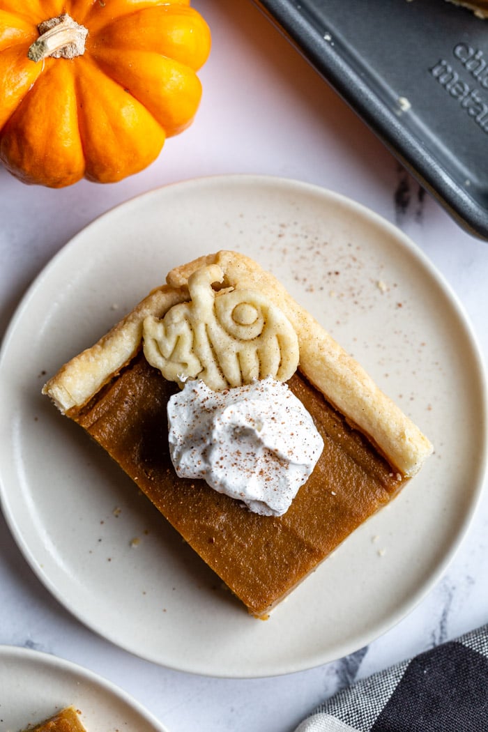 square slice of pumpkin pie topped with a pie crust pumpkin and whipped cream. a small orange pumpkin in the corner of the shot