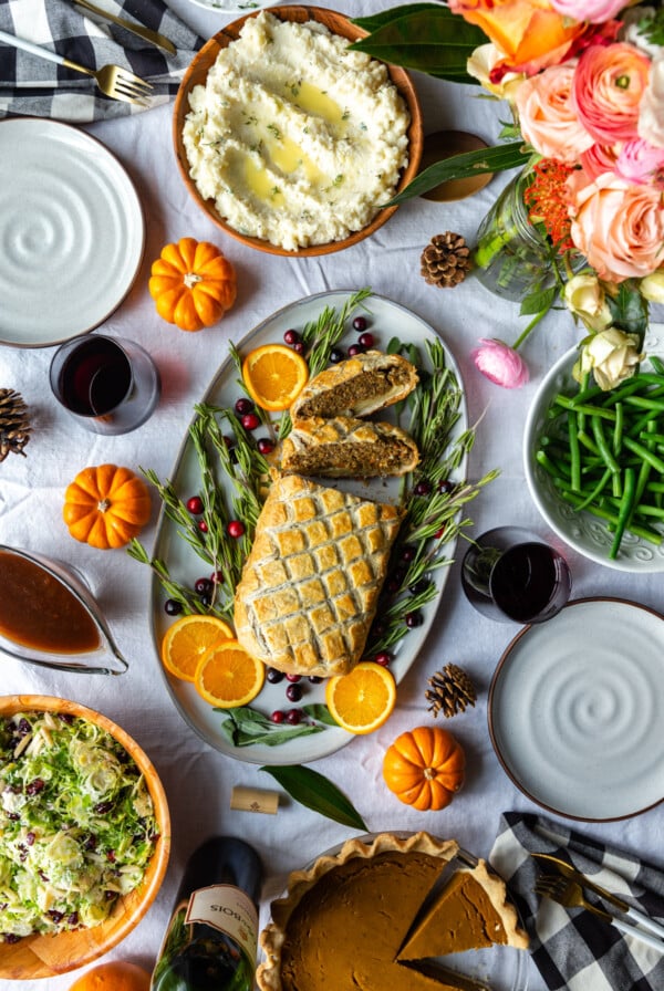 thanksgiving table filled with side dishes, a vegan wellington, wine glasses, flowers and decorations