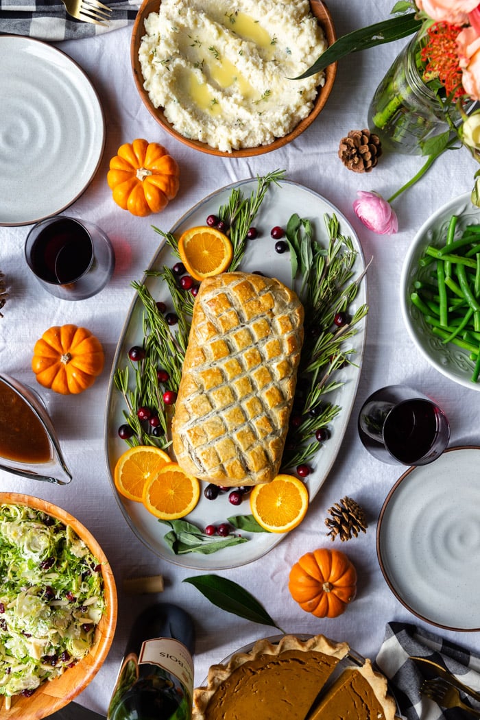 tablescape with a vegetarian wellington on a large platter surrounded by herbs and fresh fruit. Bowl of mashed potatoes and flowers on the side as well as bowls of green beans and salad