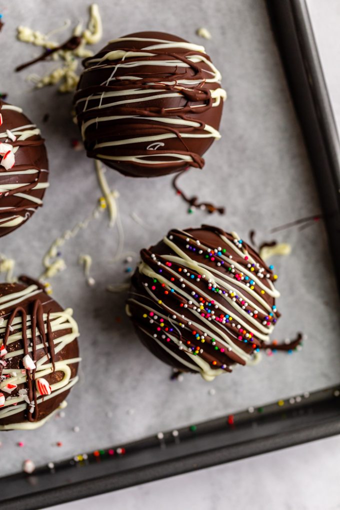 hot chocolate bombs topped with drizzled chocolate and sprinkles/ crushed peppermint all sitting on a baking sheet
