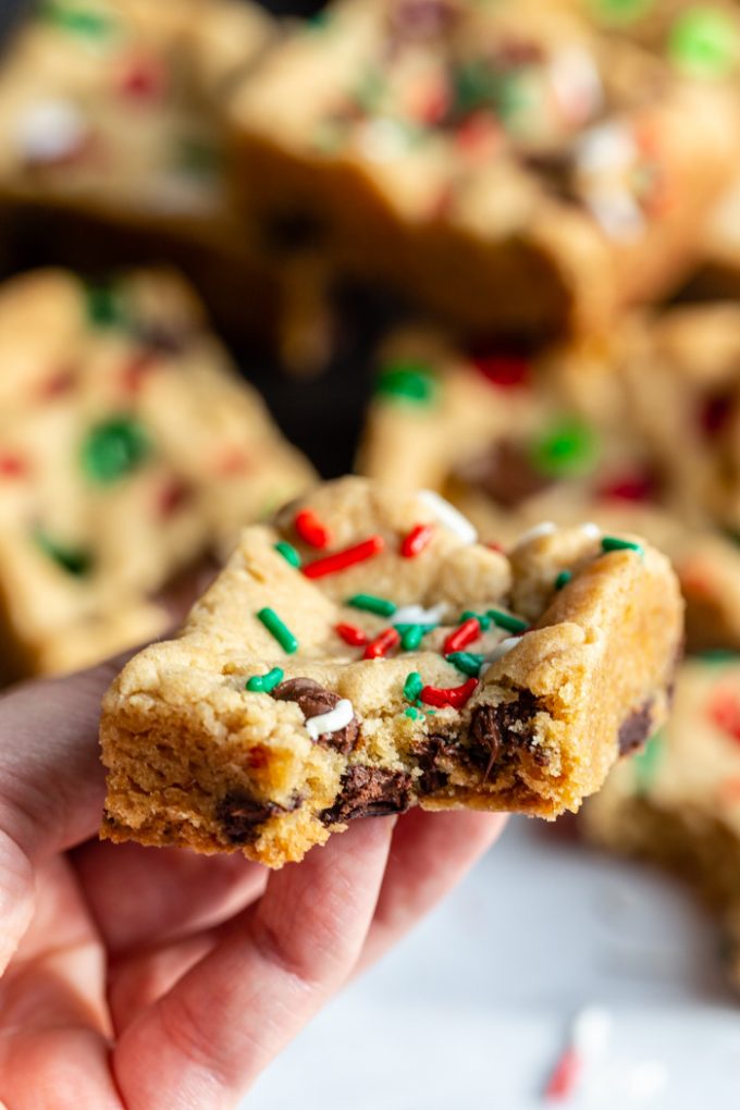 hand holding up a chocolate chip cookie bar that has red white and green sprinkles on top. more cookie bars in the background