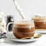 vegan hot chocolate in a clear glass with a stick of marshmallows on top of them. more hot chocolate in the background