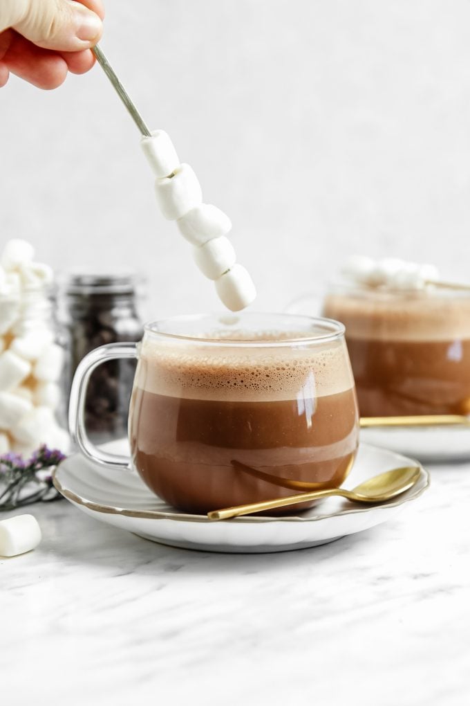 vegan hot chocolate in a clear glass with a stick of marshmallows on top of them. more hot chocolate in the background