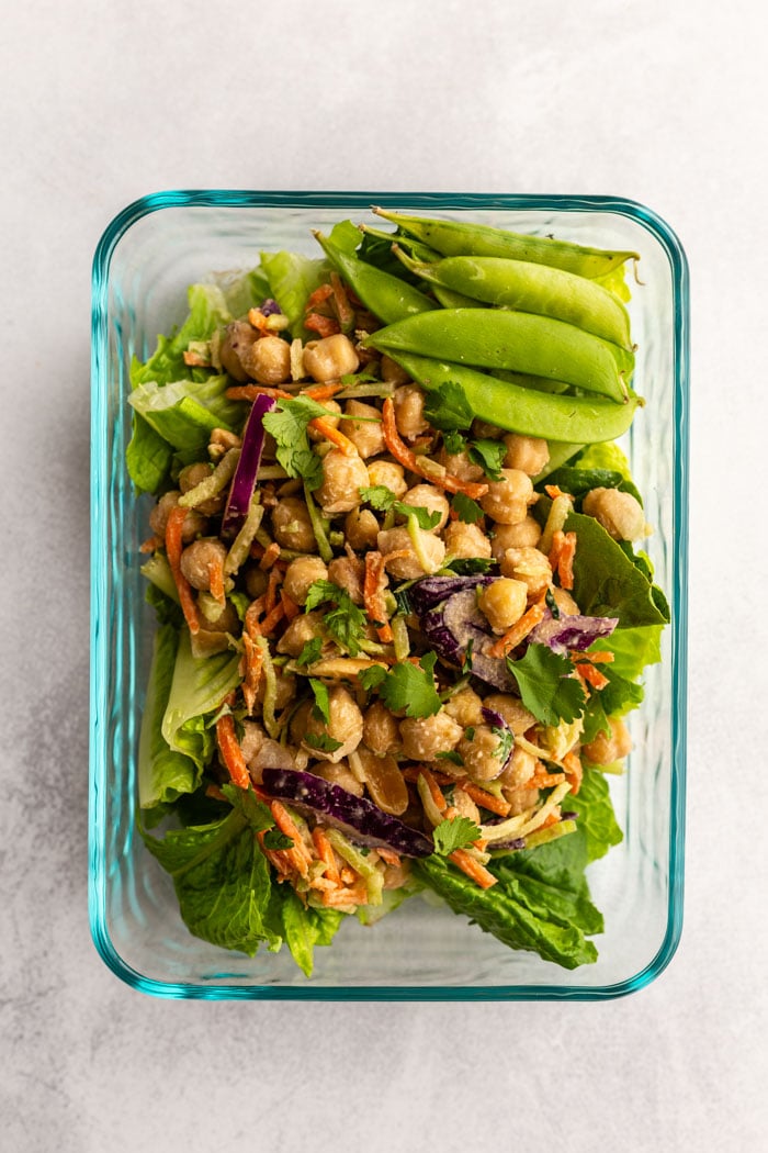 chickpea and coleslaw mix mixed together and placed atop a bed of lettuce in a meal prep container. Some sugar snap peas in the side