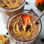 2 glasses filled with a chocolate peanut butter smoothie and topped with a slice of strawberry, shredded chocolate and a drizzle of peanut butter