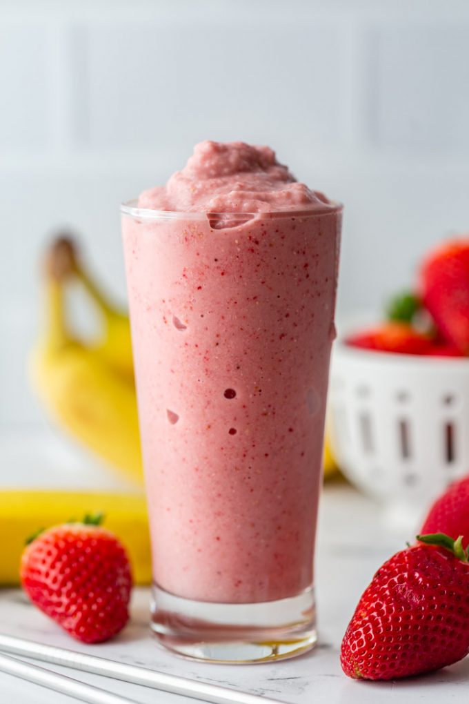 tall glass filled with a bright pink strawberry banana smoothie with fresh strawberries and bananas in the background