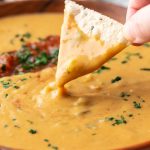 large wooden bowl filled with vegan salsa con queso and topped with salsa and cilantro. a chip scooping out some of the queso