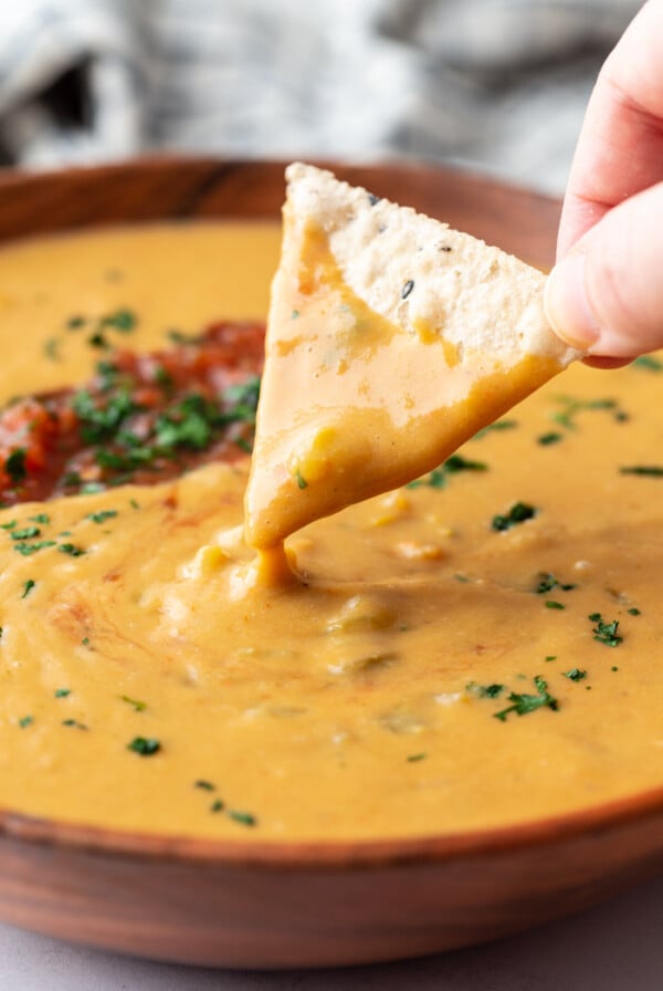 large wooden bowl filled with vegan salsa con queso and topped with salsa and cilantro. a chip scooping out some of the queso