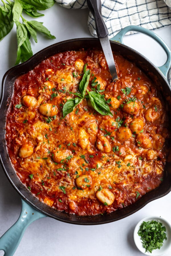 skillet with tomato sauce and gnocchi covered with cheese and garnished with fresh basil. spoon sticking out the side
