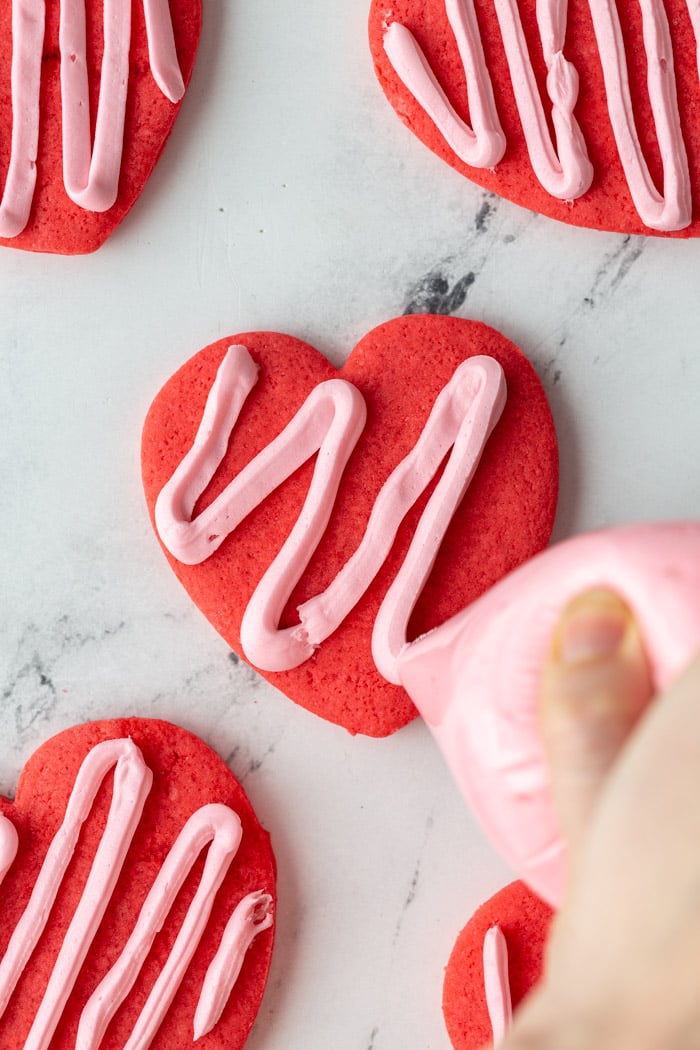 red heart cookies topped with a light pink zig zag frosting. the middle cookie is being frosted with a piping bag
