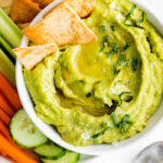 white bowl filled with green avocado hummus and surrounded by chips and veggies. a few chips sticking out of the hummus