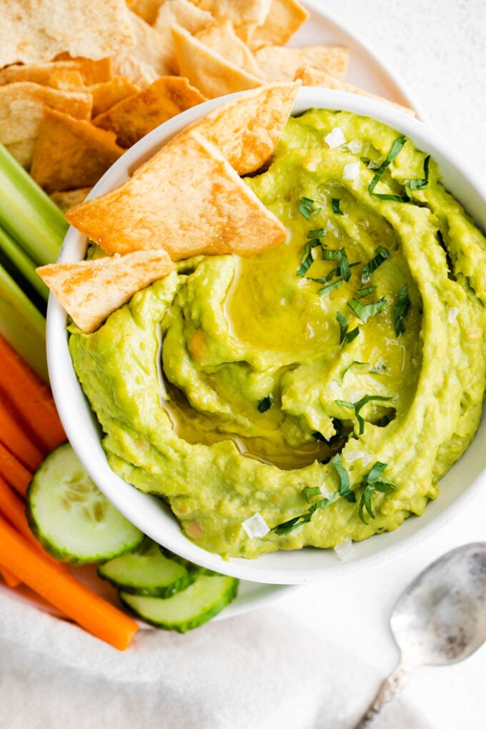 white bowl filled with green avocado hummus and surrounded by chips and veggies. a few chips sticking out of the hummus