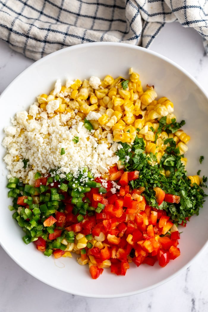 ingredients for mexican street corn salad in a large bowl including grilled corn, cotija cheese, cilantro, bell pepper, and jalapeno