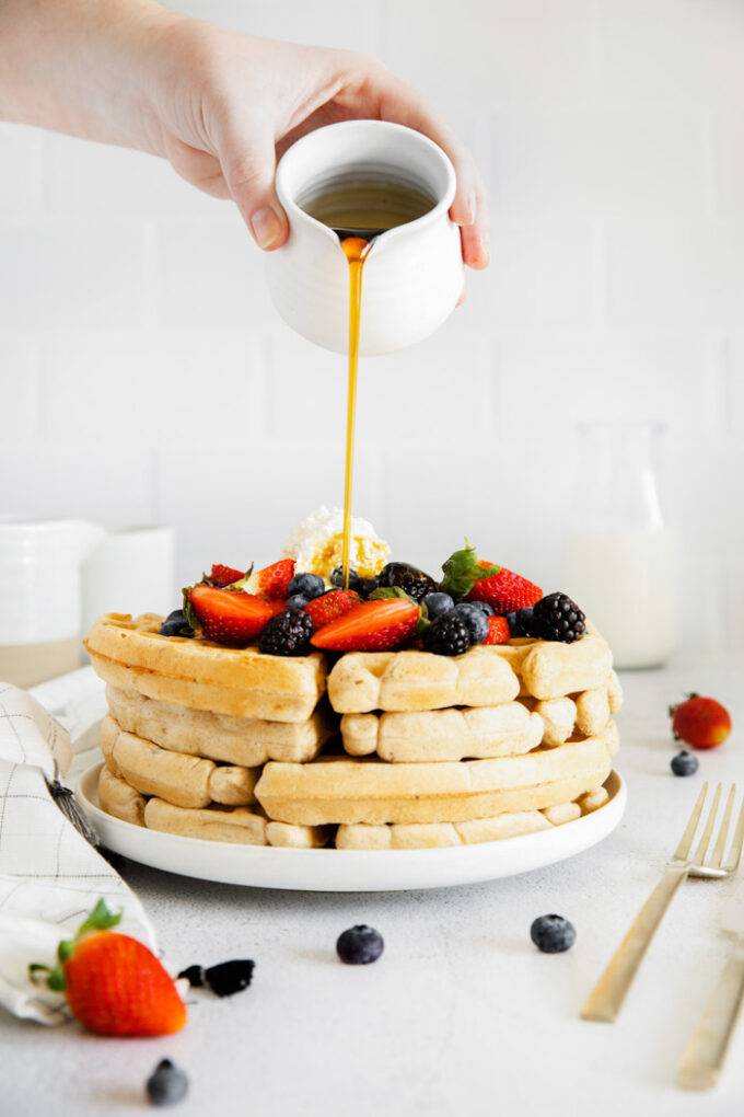 pouring maple syrup over a stack of fluffy vegan waffles