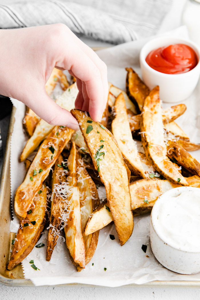 crispy potato wedges on a parchment lined baking sheet and sprinkled with cheese and herbs. ketchup and ranch on the tray. hand holding up one potato wedge