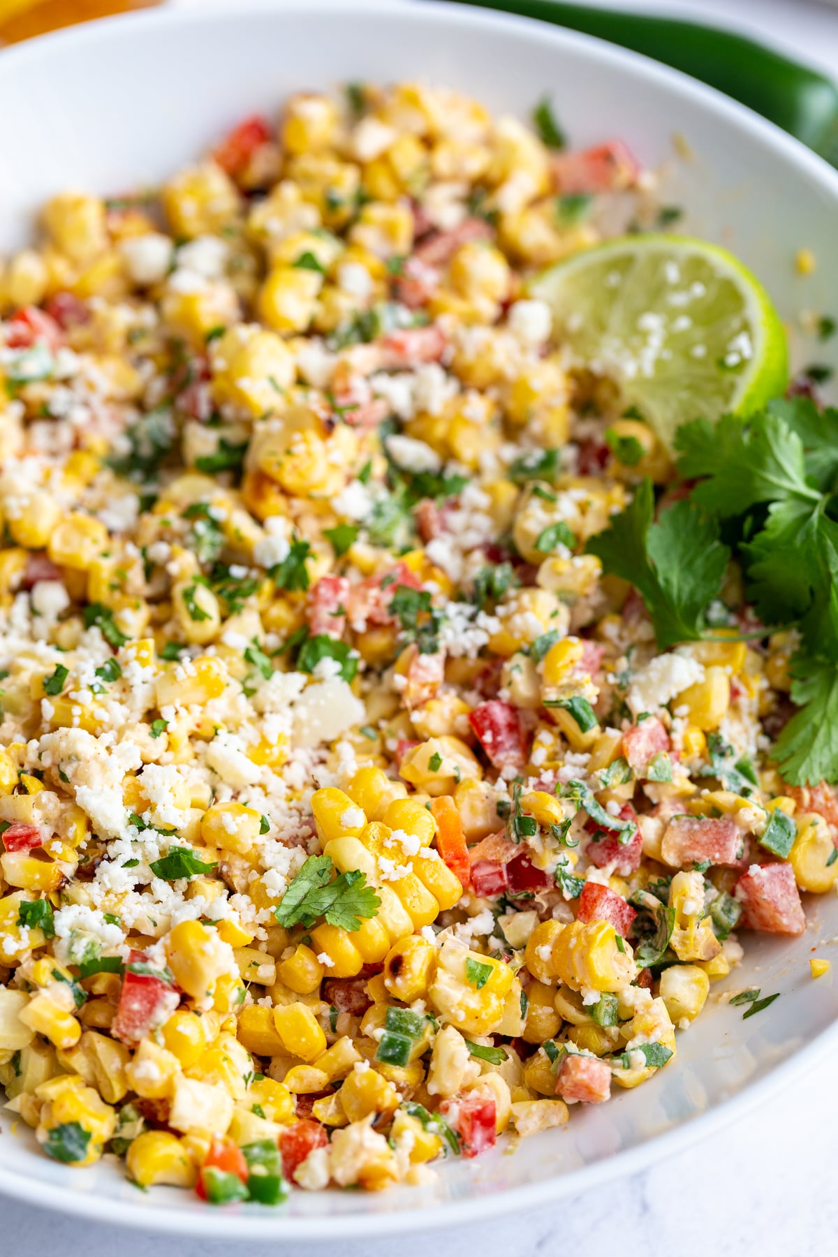 large white bowl of mexican street corn salad topped with cotija cheese and a lime with cilantro in the scene
