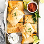 sheet pan quesadillas cut into squares and stacked on top of each other on a sheet pan with sour cream, salsa, and limes in the shot
