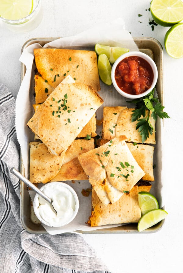 sheet pan quesadillas cut into squares and stacked on top of each other on a sheet pan with sour cream, salsa, and limes in the shot
