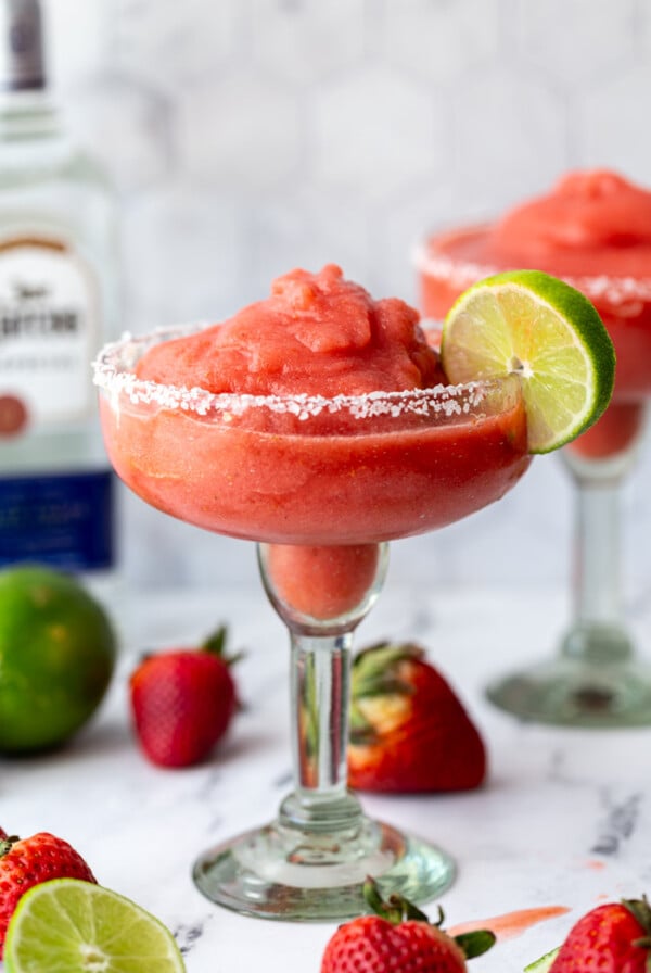 margarita glass filled with a red strawberry frozen margarita and a lime on the rim. more fruit and tequila bottle in the background