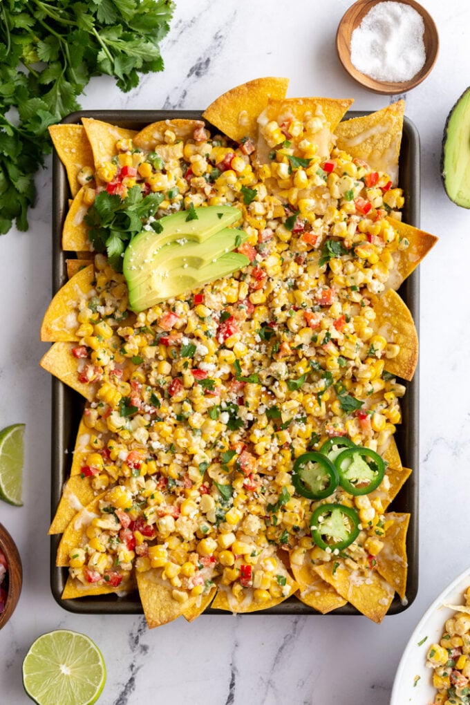 baking tray filled with nachos covered in mexican street cron. cilantro and limes in the corn of the shot