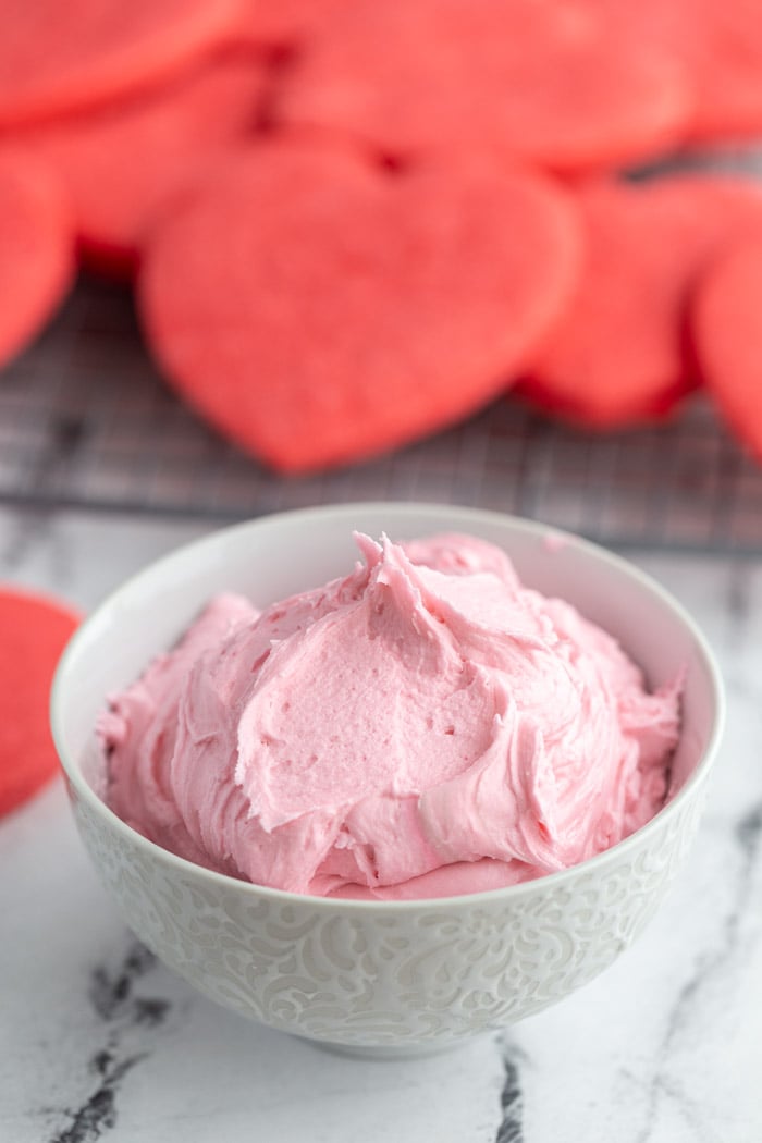 sugar cookie frosting recipe that's pink and in a small white bowl. Red cookies all around the bowl