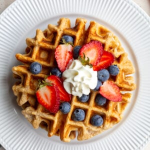 Waffle on a white plate topped with berries and whipped cream.